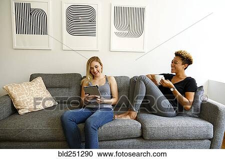 Stock Image - Women drinking coffee and using digital tablet on sofa. Fotosearch