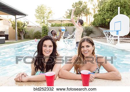 Stock Image - Women posing in swimming pool with men playing basketball. Fotosearch