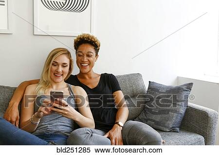 Stock Photography - Women sitting on sofa texting on cell phone. Fotosearch