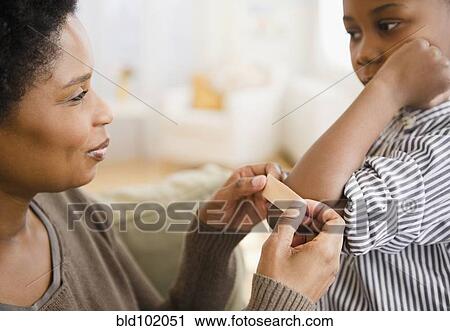 Stock Image - Black mother putting bandage on son's elbow. Fotosearch