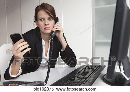 Stock Photography - Businesswoman holding cell phone and talking on landline phone. Fotosearch