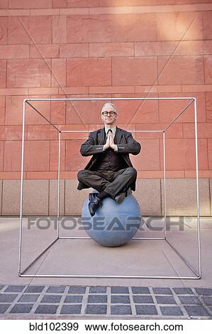 Stock Photo - Caucasian businessman practicing yoga on ball inside of box. Fotosearch