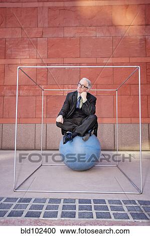 Stock Image - Caucasian businessman sitting on ball inside box. Fotosearch