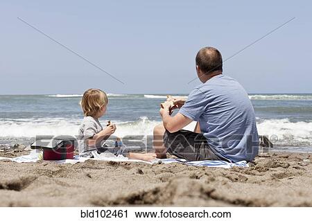 Stock Image - Caucasian father and son eating on beach. Fotosearch