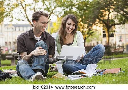 Picture - Caucasian students sitting in grass looking at laptop. Fotosearch