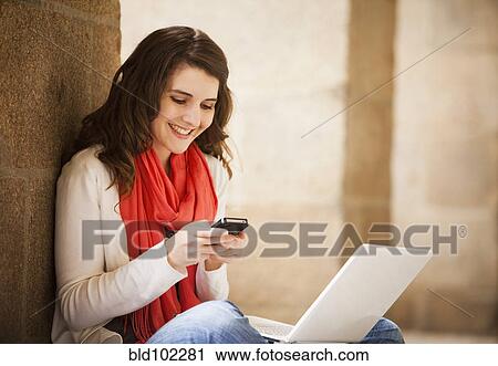 Stock Image - Caucasian woman sitting with laptop and text messaging on cell phone. Fotosearch
