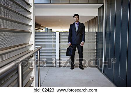Stock Image - Hispanic businessman holding briefcase in office hallway. Fotosearch