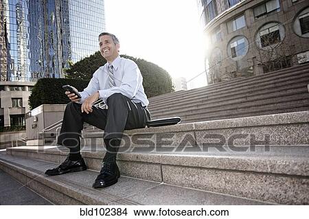 Picture - Hispanic businessman sitting on steps holding cell phone. Fotosearch