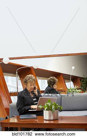 Stock Image - Hispanic businesswoman using laptop and cell phone on sofa. Fotosearch