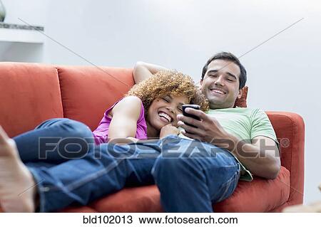 Stock Image - Mixed race couple laying on sofa looking at cell phone. Fotosearch