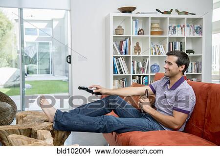Picture - Mixed race man sitting on sofa watching television. Fotosearch