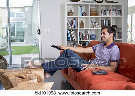Stock Photo - Mixed race man sitting on sofa watching television. Fotosearch