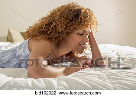 Stock Image - Mixed race woman laying on bed using digital tablet. Fotosearch