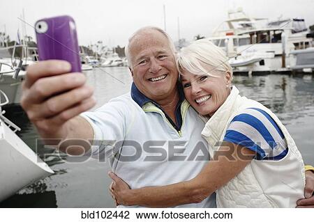 Stock Image - Smiling couple taking self-portrait at marina. Fotosearch