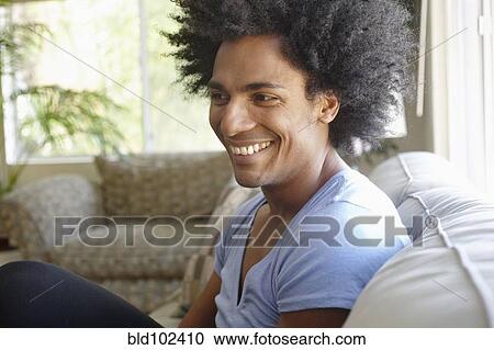 Stock Image - Smiling man sitting on sofa in living room. Fotosearch