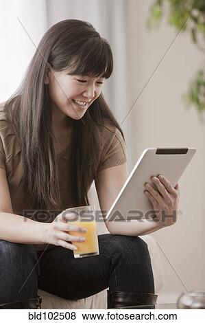 Stock Photo - Asian woman drinking orange juice and using digital tablet. Fotosearch