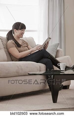 Stock Image - Asian woman sitting on sofa using digital tablet. Fotosearch