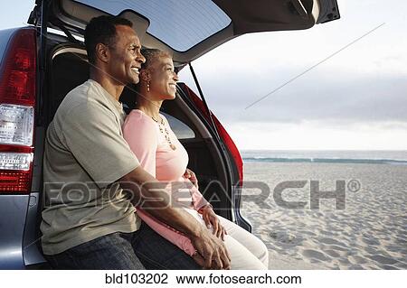 Stock Image - Black couple sitting on hatchback enjoying the beach. Fotosearch