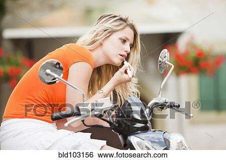 Stock Photograph - Caucasian teenager sitting on scooter putting on lipstick. Fotosearch