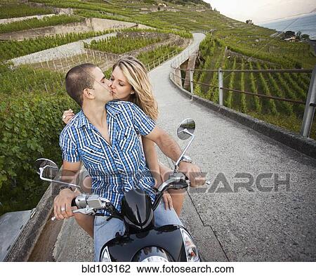 Stock Image - Couple sitting on scooter kissing in vineyard. Fotosearch