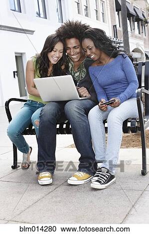 Stock Image - Friends sitting on bench using laptop. Fotosearch
