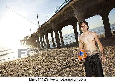 Stock Image - Hispanic man holding soccer ball on beach. Fotosearch