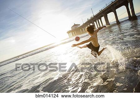 Picture - Hispanic man splashing water and kicking soccer ball on beach. Fotosearch
