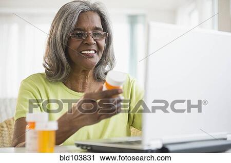 Stock Image - African American woman holding pill bottle and using laptop. Fotosearch