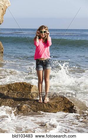 Stock Image - Caucasian teenager standing on rock in ocean. Fotosearch