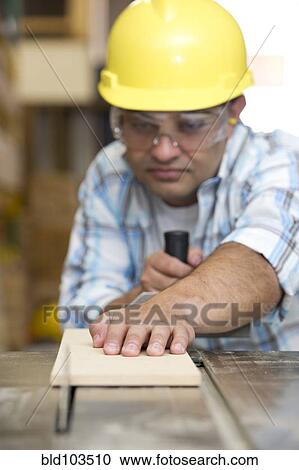 Stock Image - Hispanic carpenter cutting wood with table saw. Fotosearch