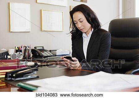 Stock Image - Mixed race businesswoman using cell phone at desk. Fotosearch