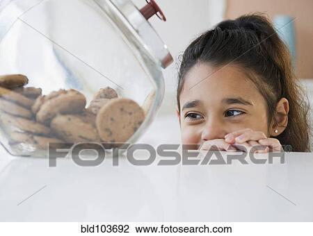 Stock Image - Mixed race girl looking at cookies in cookie jar. Fotosearch
