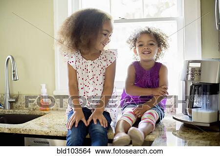 Picture - Mixed race sisters sitting on kitchen counter. Fotosearch