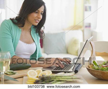Stock Photo - Mixed race woman using laptop in kitchen. Fotosearch