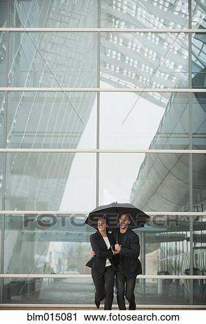 Stock Image - Caucasian business people running together underneath umbrella. Fotosearch