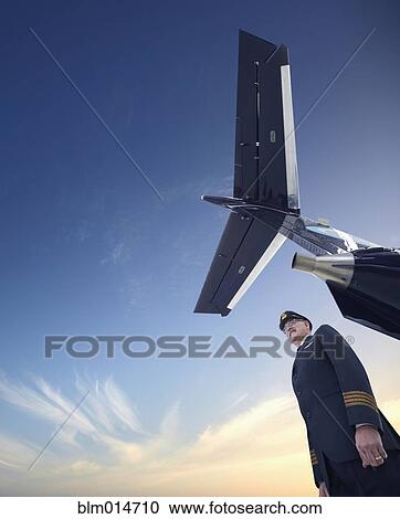 Stock Image - Caucasian pilot standing underneath airplane. Fotosearch