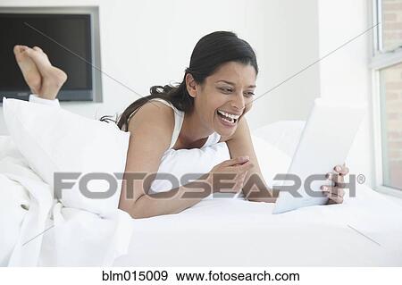 Stock Photo - Laughing Hispanic woman laying in bed using digital tablet. Fotosearch
