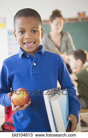 Stock Image - African American boy holding apple in classroom. Fotosearch