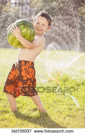 Stock Photo - Caucasian boy carrying watermelon through sprinkler. Fotosearch