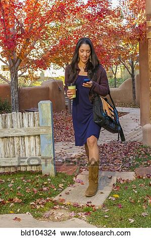 Picture - Hispanic woman carrying coffee and text messaging on cell phone. Fotosearch