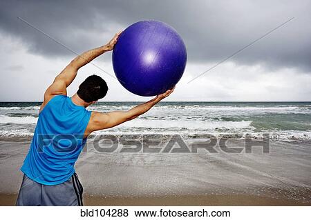 Stock Photo - Young man holding exercise ball on beach. Fotosearch