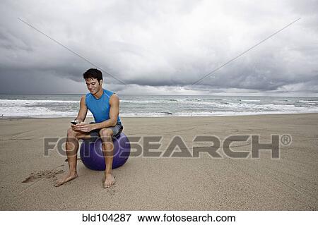 Stock Photo - Young man sitting beach text messaging on cell phone. Fotosearch