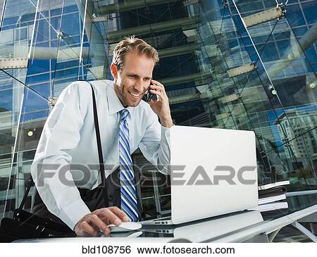 Stock Photograph - Caucasian businessman talking on cell phone at laptop. Fotosearch