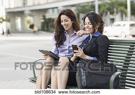 Hispanic businesswomen on bench using cell phone and digital tablet View Large Photo Image Picture - Hispanic businesswomen on bench using cell phone and digital tablet. Fotosearch