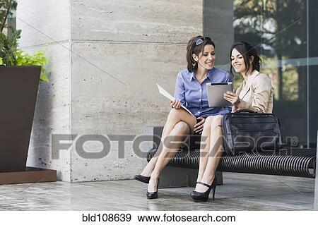 Stock Photo - Hispanic businesswomen sitting on bench looking at digital tablet. Fotosearch