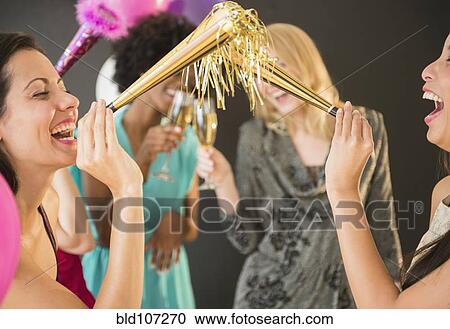 Stock Image - Smiling women blowing party horns at party. Fotosearch