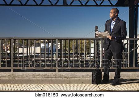 Stock Image - Black businessman reading newspaper and waiting at train station. Fotosearch