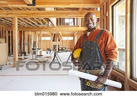 Stock Image - Black construction worker holding blueprints in unfinished room. Fotosearch