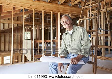 Stock Image - Caucasian construction worker holding blueprints in unfinished room. Fotosearch