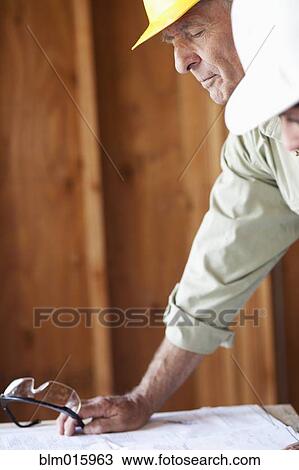 Stock Image - Caucasian construction worker looking at blueprints in unfinished room. Fotosearch